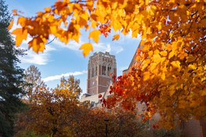 A tall brick clock tower rises behind trees with bright orange and red autumn leaves under a partly cloudy sky.