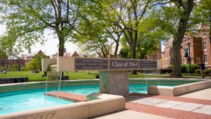 A concrete fountain with water flowing from a rectangular top labeled Class of 1964 sits among green trees and brick.