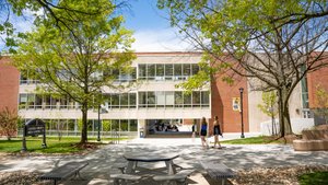 Two women walk on a paved path past benches and trees toward a modern building with large windows.