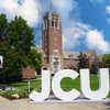 Large white letters spelling JCU stand on a sidewalk in front of a tall brick clock tower and green lawn under a partly.