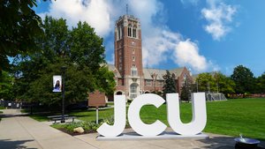 Large white letters spelling JCU stand on a sidewalk in front of a tall brick clock tower and green lawn under a partly.