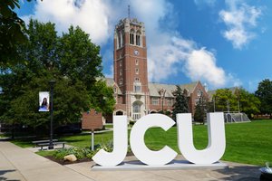 Large white letters spelling JCU stand on a sidewalk in front of a tall brick clock tower and green lawn under a partly.