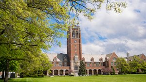 A large brick building with a tall clock tower stands behind a green lawn framed by leafy trees under a partly cloudy sky.