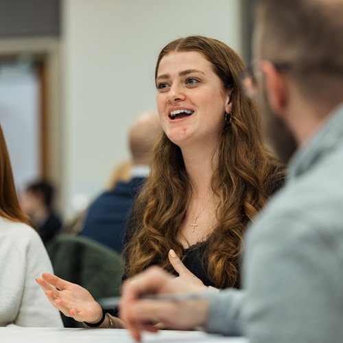 A young woman with long curly hair gestures and smiles while talking at a table with others indoors.