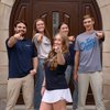 Five smiling young adults wearing John Carroll University shirts point toward the camera in front of a large wooden door.