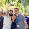 Three people smiling and posing for a selfie under green tree branches on a sunny day at John Carroll University.