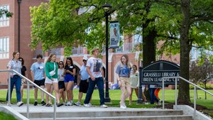 A group of people walk down steps near a sign for Grasselli Library and Breen Learning Center on a green campus.