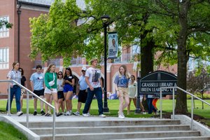 A group of people walk down steps near a sign for Grasselli Library and Breen Learning Center on a green campus.