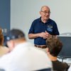 An older man wearing glasses and a navy polo shirt with a Boler College of Business name tag speaks to a seated audience.