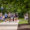 A group of people walk along a tree-lined sidewalk on a campus at John Carroll University on a cloudy day.