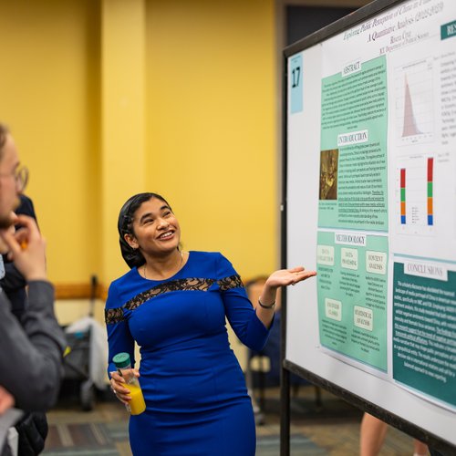 A woman in a blue dress holds a bottle and gestures toward a research poster while a man listens attentively.