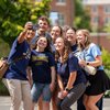 A group of six people wearing John Carroll University shirts smile as one person takes a selfie outdoors on campus.