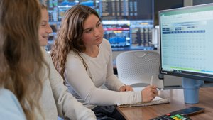 Two students study financial data on a computer screen in a room with multiple monitors displaying charts.