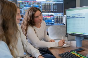 Two students study financial data on a computer screen in a room with multiple monitors displaying charts.