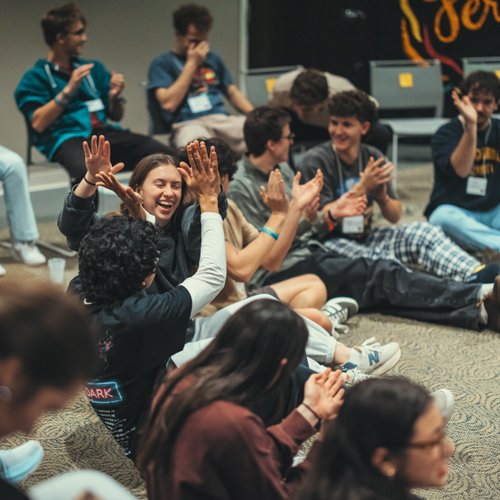 Students laugh and clap together, some sitting on chairs, and others cross-legged on the floor.
