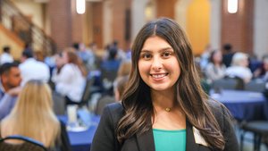 A young woman with long dark hair smiles at the camera in a busy indoor space with people seated at tables behind her.
