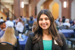A young woman with long dark hair smiles at the camera in a busy indoor space with people seated at tables behind her.
