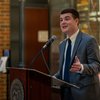 A man in a gray suit and blue tie speaks at a podium with a John Carroll University seal indoors.