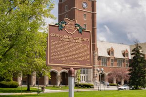 A brown historical marker for John Carroll University, founded in eighteen eighty-six, stands on a sunny campus lawn near