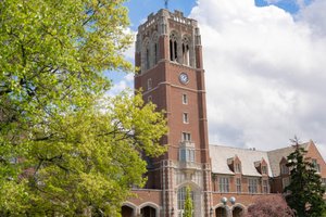 View of a tall brick clock tower and adjacent building framed by green trees and lawn under a partly cloudy blue sky.