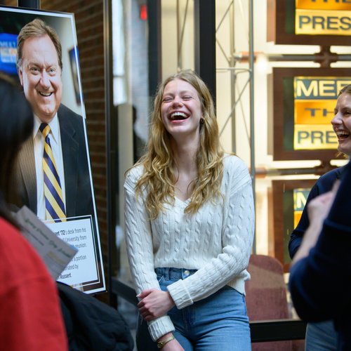 A young woman in a white sweater laughs with two other people near a John Carroll University poster of a man in a suit.