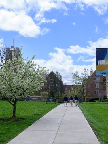 View of a concrete pathway framed by green lawns and blossoming trees leading to brick campus buildings at John Carroll.