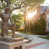 A bronze statue of a kneeling armored man holds a sword on the grassy quad, sunlight dappling through trees.