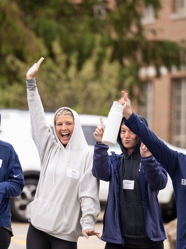 Four young women wearing hoodies and name tags raise their arms and smile outside a brick campus building at John Carroll.