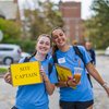 Two smiling young women wearing blue Jesuit Day of Service shirts hold yellow "Site Captain" signs on a campus street.