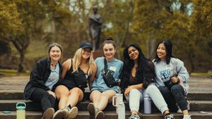 Five students sit on stone steps outside on a campus path framed by trees and a statue in the background.
