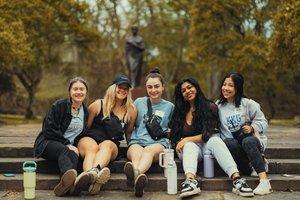 Five students sit on stone steps outside on a campus path framed by trees and a statue in the background.