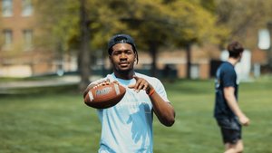 A young man wearing a light blue shirt and backward cap holds a football on a grassy area at John Carroll University.