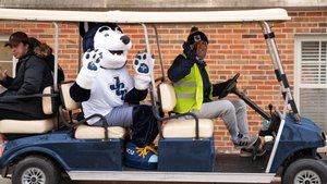 A husky mascot and two students wave from a blue golf cart on JCU campus; the driver wears a reflective vest.