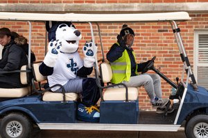 A husky mascot and two students wave from a blue golf cart on JCU campus; the driver wears a reflective vest.