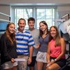 Five people smile together inside a dorm room with bunk beds and a window with blinds behind them.