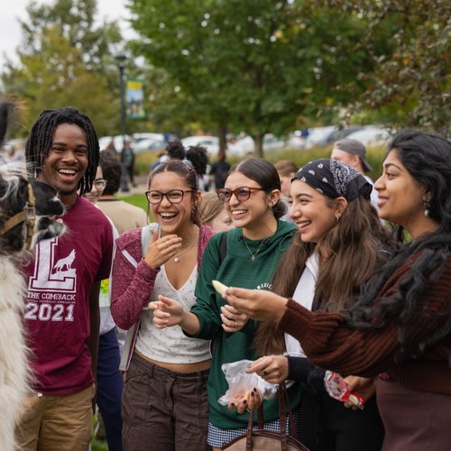 A group of five smiling students interact with a llama on a grassy area framed by trees at John Carroll University.