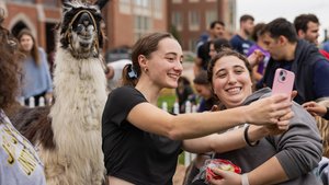 Two young women smile as they take a selfie with a llama on a John Carroll University campus lawn.