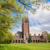 View of a tall brick clock tower and adjacent campus building framed by green trees and a large grassy lawn under a partly.