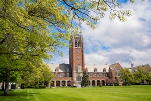 View of a tall brick clock tower and adjacent campus building framed by green trees and a large grassy lawn under a partly.