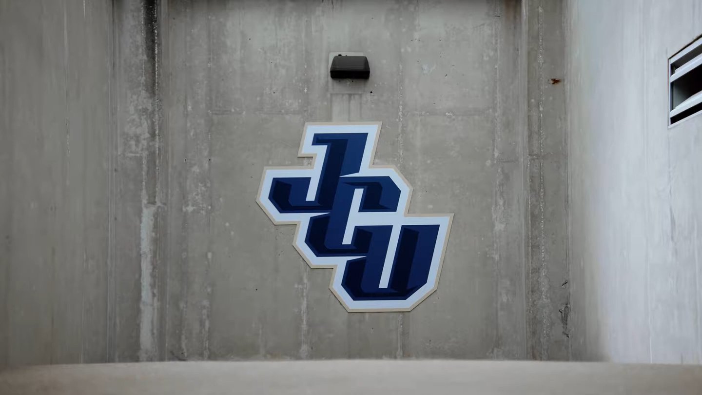 Blue and white JCU letters logo mounted on a gray concrete wall with a black light fixture above it.
