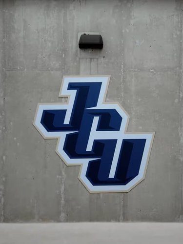 Blue and white JCU letters logo mounted on a gray concrete wall with a black light fixture above it.