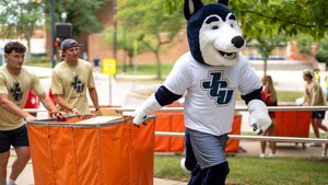 The John Carroll University mascot in a husky costume pulls an orange cart with two men in matching JCU football shirts.