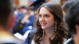 A student with long brown hair and heart-shaped earrings smiles brightly during a graduation ceremony on the JCU campus.
