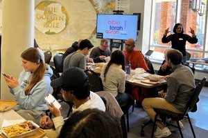 A group of people sit around tables eating and talking while a woman stands near a screen showing religious texts.