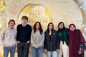 Six young adults stand smiling in front of a mural depicting symbols and names of various world religions.
