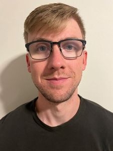 A young man with light brown hair and glasses looks directly at the camera with a slight smile against a plain wall.