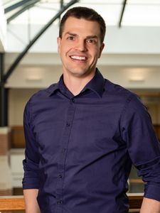 A man in a dark purple shirt smiles while leaning on a wooden railing inside a bright modern building.