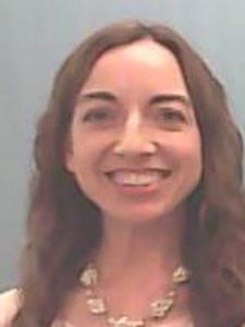 A woman with long brown hair smiles at the camera against a plain light gray background.