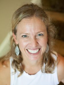 A woman with curly blonde hair smiles widely at the camera wearing a white sleeveless top and silver earrings.