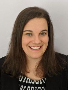 A woman with straight brown hair smiles at the camera wearing a black jacket and a patterned top.